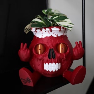 May include: A red skull-shaped plant pot with a white rim and dripping red details. The pot has orange eyes, a black nose, and white teeth. It contains a green and white plant, and is making a peace sign with its hands.