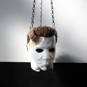 May include: A white mask with brown hair and a partially exposed face, suspended by black chains. The mask has dark eye holes and a detailed facial structure, with a blend of white and flesh-toned paint. The chains hang against a white and black background.