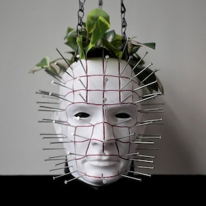 May include: A white, human-shaped planter featuring a grid of red lines across the face, with numerous silver metal nails protruding from the surface. A green leafy plant grows from the top, and the planter is suspended by black metal chains.