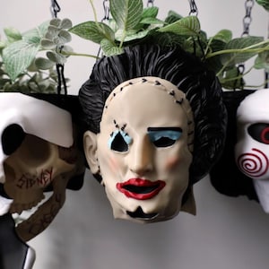 May include: A hanging planter set featuring three horror-themed heads. The centre head wears a stitched mask with red lips and blue eyeshadow. To the left is a skull with the name Sidney written in red, and to the right is a white puppet mask.