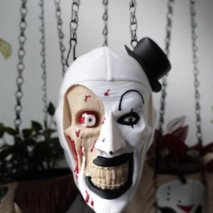 May include: A Halloween hanging mask with a clown design, half skull appearance. The mask is white with black accents, a small black top hat, and red blood-like details. Other masks are visible in the background.