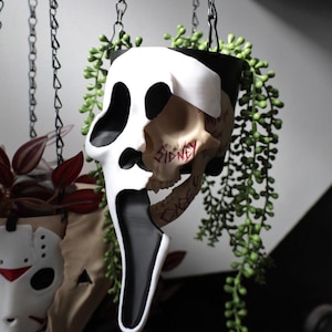 May include: A Scream mask-shaped hanging planter, featuring a skull design with the word "SIDNEY" inscribed. The planter is black and white, with trailing green plants. Another mask is visible in the background.