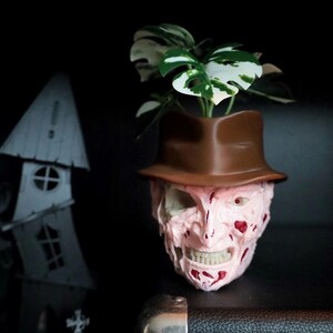 May include: A novelty planter in the shape of a horror movie character's face, with a brown hat. The pink and red face holds a green and white plant. A spooky house and graveyard are in the background.