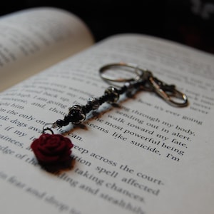 May include: A silver keychain with a beaded stem and a dark red rose charm rests on an open book. The book's pages display printed text. The keychain's design includes small, dark beads and a rose.