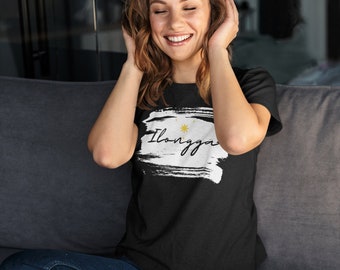 May include: A woman wearing a black t-shirt with a white design that says "Ilongga" with a yellow sun and stars. She is wearing blue jeans and has headphones on.