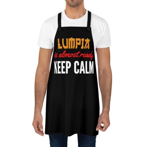 Puede incluir: Delantal negro con texto blanco que dice "LUMPIA is almost ready KEEP CALM". El texto est&aacute; en amarillo y naranja con acentos rojos.