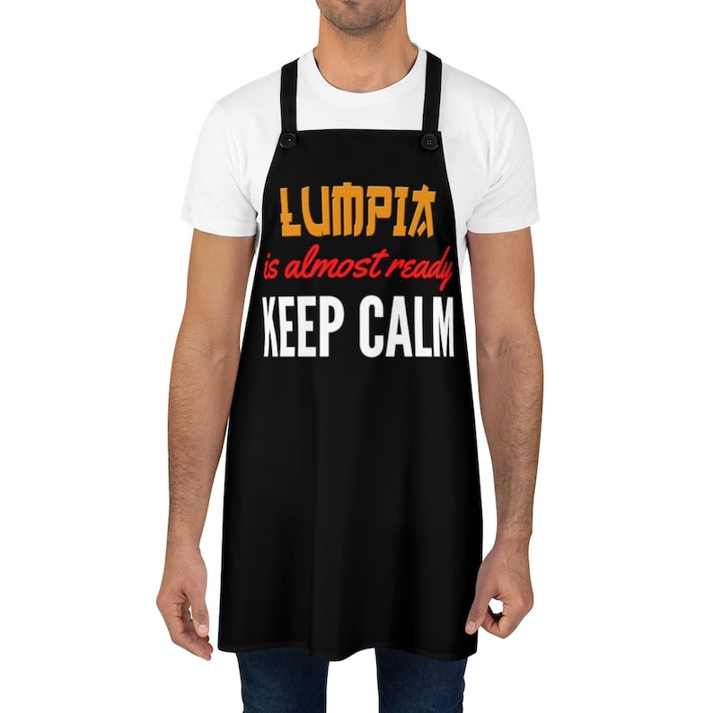 Puede incluir: Delantal negro con texto blanco que dice "LUMPIA is almost ready KEEP CALM". El texto est&aacute; en amarillo y naranja con acentos rojos.