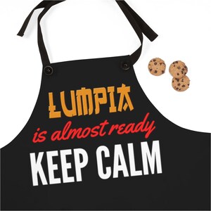 Puede incluir: Delantal negro con texto blanco que dice "LUMPIA is almost ready KEEP CALM". El texto "LUMPIA" est&aacute; en naranja y el texto "almost ready" est&aacute; en rojo. Tres galletas con chispas de chocolate est&aacute;n en la esquina superior derecha de la imagen.