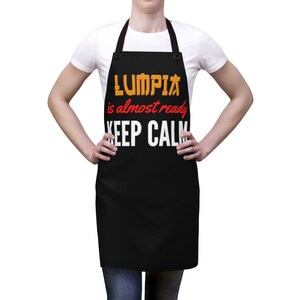 Puede incluir: Delantal negro con el texto "LUMPIA is almost ready KEEP CALM" en naranja, rojo y blanco.