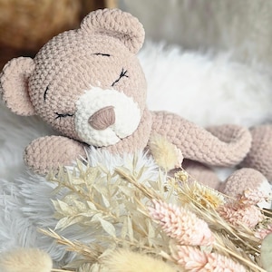Puede incluir: Un oso de peluche de crochet con los ojos cerrados y una nariz blanca, acostado sobre una superficie blanca y esponjosa con flores secas.