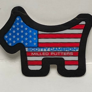 Pode incluir: Marcador de bola de golfe Scotty Cameron em forma de cão, com o design da bandeira americana. Apresenta um campo de estrelas azul e riscas vermelhas e brancas, uma borda preta e o texto "SCOTTY CAMERON MILLED PUTTERS".