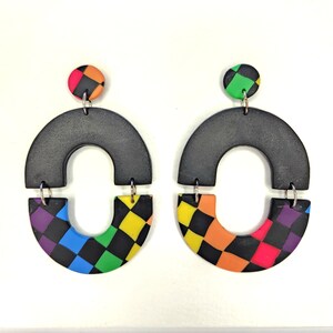 May include: A pair of black and rainbow checkerboard patterned statement earrings. Each earring features a black half-circle with a rainbow checkerboard half-circle attached to it. The earrings are suspended from a black circle with a rainbow checkerboard pattern.