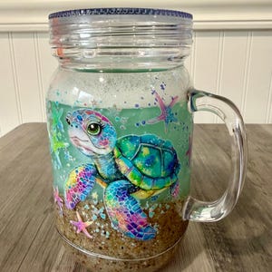 Puede incluir: Tarro de cristal transparente con asa y tapa azul. El tarro presenta un colorido diseño de tortuga marina con detalles de arcoíris y estrellas de mar. El fondo del tarro está lleno de arena y el líquido del interior es de color verde claro.