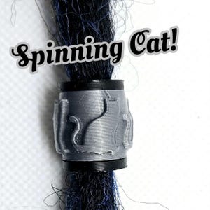 May include: A silver dreadlock bead with a spinning cat design. The bead is on a black dreadlock.