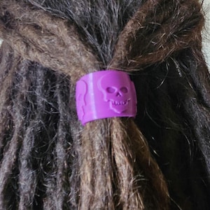 May include: A purple hair accessory featuring a skull design. The accessory is wrapped around a section of dark brown dreadlocks. The skull is a prominent element, adding a gothic or alternative aesthetic to the hairstyle.