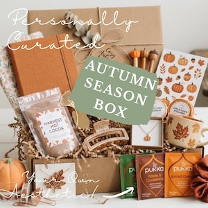 May include: A cardboard gift box filled with autumn-themed items including an orange notebook, a pouch of Harvest Hot Cocoa, a gold-toned hair claw, a pumpkin necklace, a mug with leaf patterns, Pukka tea bags, and a sheet of pumpkin stickers.