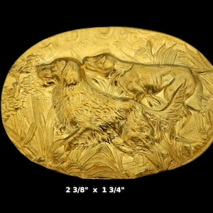 May include: A gold-toned metal belt buckle featuring a detailed relief sculpture of two dogs in a grassy field. The buckle measures 2 3/8 inches by 1 3/4 inches.