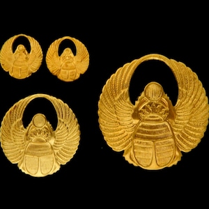 May include: Set of four gold-colored scarab beetle amulets with wings. The amulets are in various sizes and are made of a shiny metal.