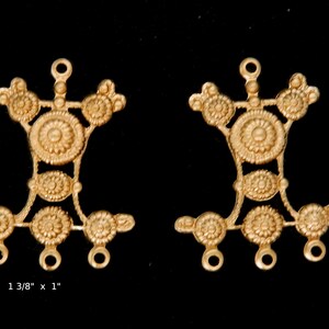 May include: Two gold-toned metal jewelry findings with a decorative design. Each finding has a circular center with a smaller circle in the center. The findings have three loops at the bottom. The findings measure 1 3/8 inches by 1 inch.