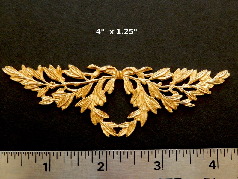 K&ouml;nnte beinhalten: Goldene Metall-Dekorverzierung mit einem Blattdesign und einer Schleife in der Mitte. Die Verzierung misst 10,16 cm x 3,17 cm.
