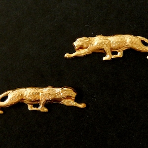 May include: Two gold-colored metal leopard figurines, each with a detailed textured surface, are posed in a crouching position on a black background.