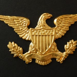 May include: A gold-colored metal emblem of an eagle with outstretched wings, a shield, and a banner. The eagle is highly detailed, with textured feathers and a prominent beak. The emblem is likely a decorative element or a symbol of authority.