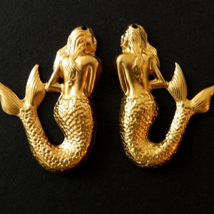 May include: Two gold-tone mermaid charms with a detailed, textured finish. The mermaids are facing to the right and have long flowing hair. Their tails are curled and have a scaly texture.