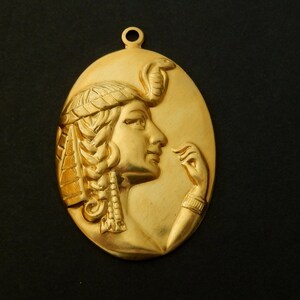 May include: A gold-colored pendant featuring a detailed relief of a woman's profile with a snake on her head. The woman is wearing a headdress and has a hand raised to her face.