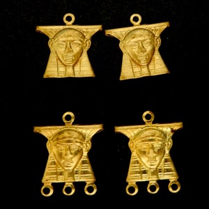 May include: Four gold-colored metal pendants featuring a raised image of a pharaoh's head. The pendants have a loop at the top for hanging and three loops at the bottom for attaching to a chain or other jewelry.