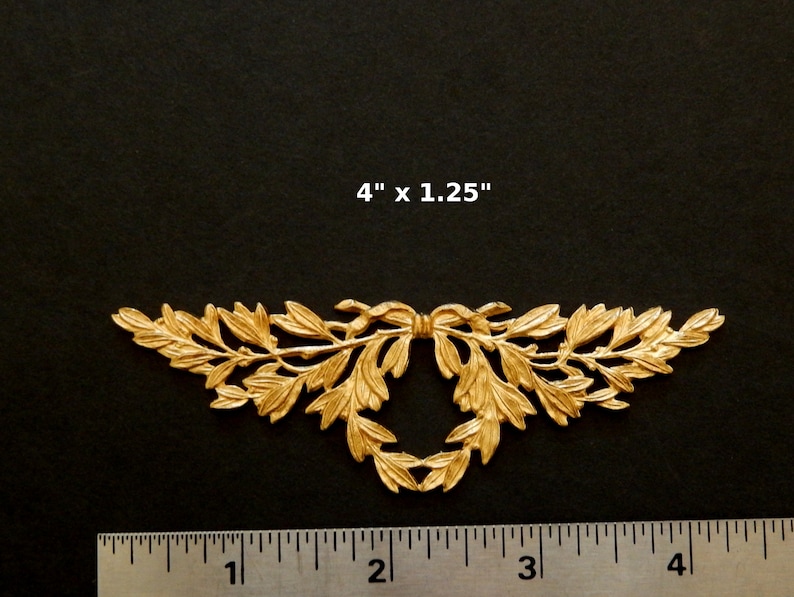 K&ouml;nnte beinhalten: Goldene Metall-Dekorverzierung mit einem Blattdesign und einer Schleife in der Mitte. Die Verzierung misst 10,16 cm x 3,18 cm.
