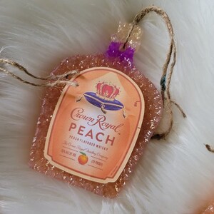 Op de afbeelding: Een ornament in de vorm van een Crown Royal whiskyfles met perzik smaak. Het ornament is gemaakt van helder hars met een glinsterende afwerking en heeft een perzikkleurig etiket met het Crown Royal logo en de woorden "Perzik Smaak Whisky".