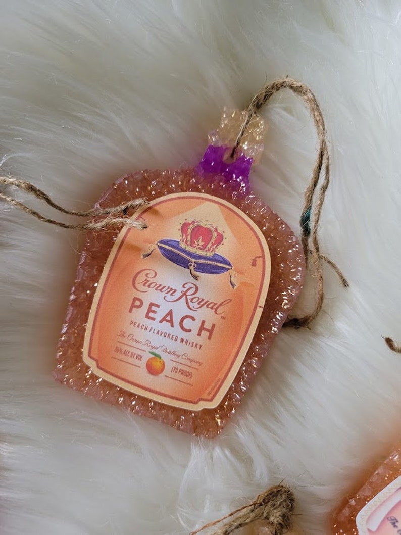 Op de afbeelding: Een ornament in de vorm van een Crown Royal whiskyfles met perzik smaak. Het ornament is gemaakt van helder hars met een glinsterende afwerking en heeft een perzikkleurig etiket met het Crown Royal logo en de woorden "Perzik Smaak Whisky".
