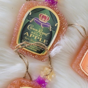 Op de afbeelding: Een roze en oranje ornament in de vorm van een Crown Royal Regal Apple likeurfles met een gouden en paarse kroon op het etiket. Het ornament is gemaakt van een getextureerd materiaal en heeft een touwtje om op te hangen.