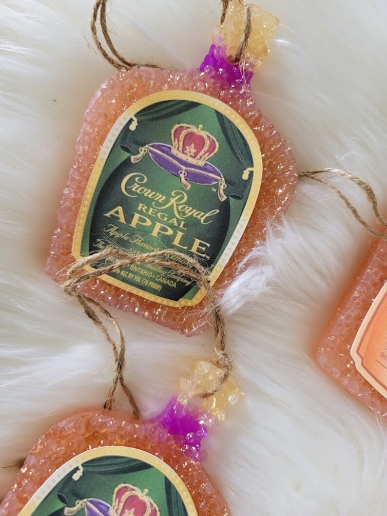 Op de afbeelding: Een roze en oranje ornament in de vorm van een Crown Royal Regal Apple likeurfles met een gouden en paarse kroon op het etiket. Het ornament is gemaakt van een getextureerd materiaal en heeft een touwtje om op te hangen.