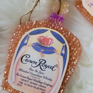 Op de afbeelding: Een perzikkleurige autoluchtverfrisser in de vorm van een Crown Royal-likeurfles. De fles heeft een gouden kroon en een blauw en wit etiket met de tekst "Crown Royal Fine De Luxe Blended Canadian Whisky A Product of The Crown Royal Distilling Company Toronto Ontario Canada 750ML ALC 40% BY VOL (80 PROOF) DSP100 DIPIE".
