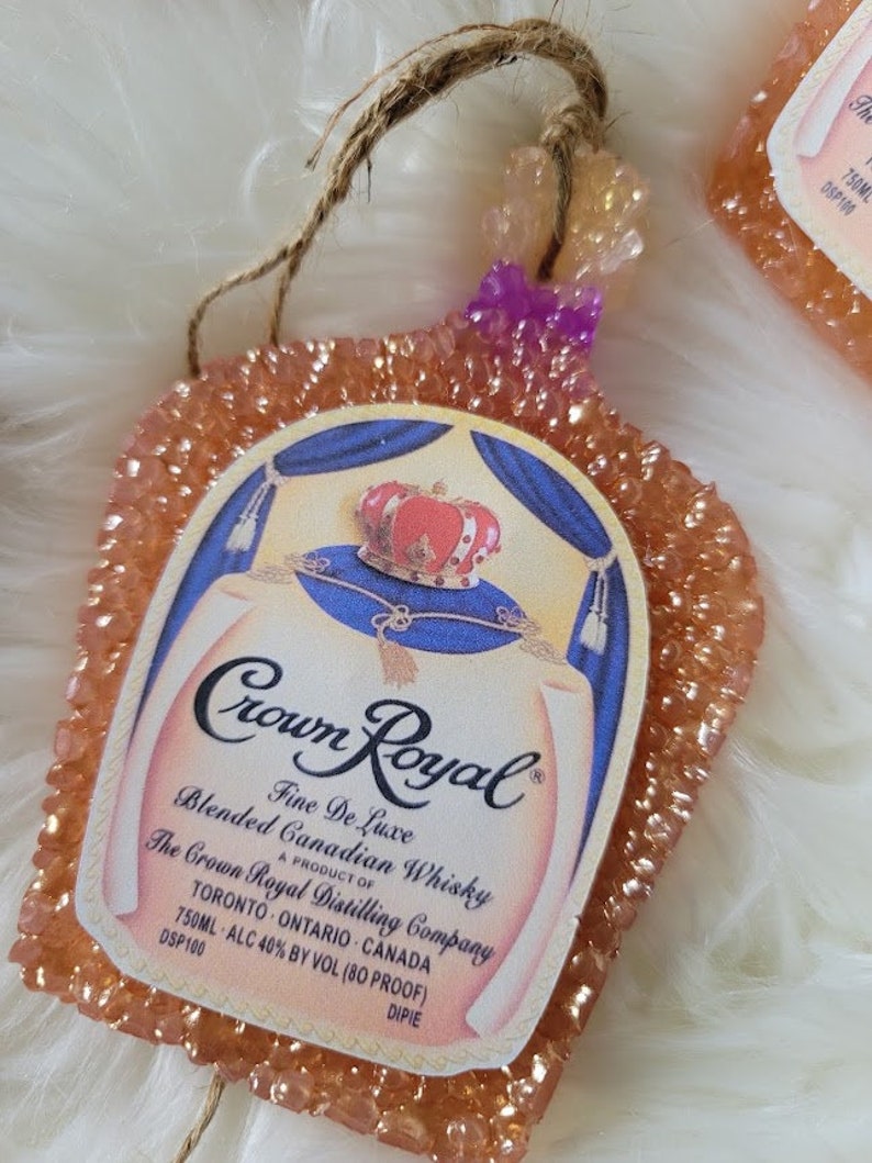 Op de afbeelding: Een perzikkleurige autoluchtverfrisser in de vorm van een Crown Royal-likeurfles. De fles heeft een gouden kroon en een blauw en wit etiket met de tekst "Crown Royal Fine De Luxe Blended Canadian Whisky A Product of The Crown Royal Distilling Company Toronto Ontario Canada 750ML ALC 40% BY VOL (80 PROOF) DSP100 DIPIE".