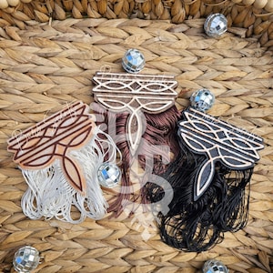 May include: Three decorative wooden ornaments with intricate designs and fringe in white, burgundy, and black. The ornaments are displayed on a woven basket, with small mirrored disco balls scattered around.