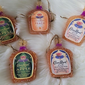 Op de afbeelding: Vijf Crown Royal likeurfles-versieringen. De versieringen zijn gemaakt van helder glas met een glinsterende afwerking. De flessen zijn versierd met het Crown Royal logo en verschillende smaken, waaronder appel, perzik en Fine De Luxe. De versieringen hangen aan een touwtje.