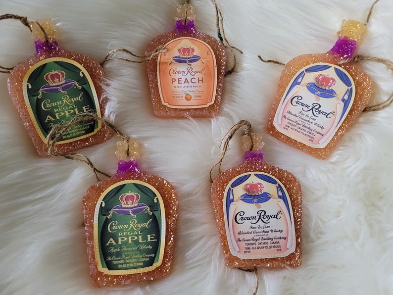 Op de afbeelding: Vijf Crown Royal likeurfles-versieringen. De versieringen zijn gemaakt van helder glas met een glinsterende afwerking. De flessen zijn versierd met het Crown Royal logo en verschillende smaken, waaronder appel, perzik en Fine De Luxe. De versieringen hangen aan een touwtje.