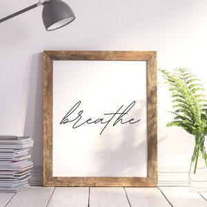 May include: A framed print with the word "breathe" in elegant cursive script. The frame is made of rustic wood. A gray desk lamp and a stack of magazines are to the left. A vase of green ferns is to the right.