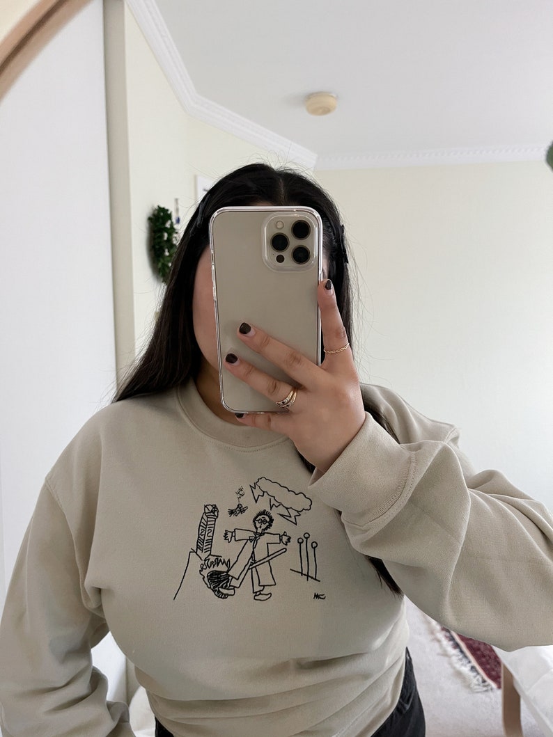 May include: A light beige sweatshirt with a black and white illustration of a stick figure holding a broom and standing in front of a building. The illustration includes the text "MC".