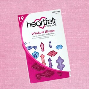 May include: A package of Heartfelt Creations Window Hinges craft dies. The package includes 19 pieces of various decorative shapes, including keys, hinges, and ornate designs. The package is white and pink with the Heartfelt Creations logo and product information.