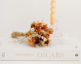 May include: A small bouquet of dried flowers in shades of orange, brown, and white, tied with twine. The bouquet is sitting on a white book with the text "RED CARPET OSCARS" printed on the cover. A tall, twisted, peach-colored candle is in the background.