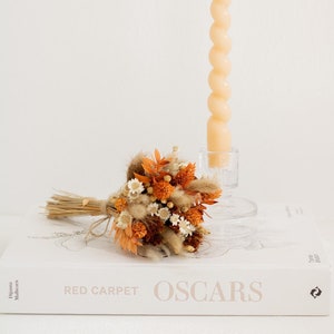 Könnte beinhalten: Ein kleiner Strauß getrockneter Blumen in Orange-, Braun- und Weißtönen, mit Schnur gebunden. Der Strauß liegt auf einem weißen Buch mit dem aufgedruckten Text "RED CARPET OSCARS". Im Hintergrund steht eine hohe, gedrehte, pfirsichfarbene Kerze.