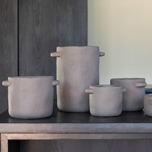 Può includere: Quattro vasi in ceramica grigia con manici, disposti in fila su una superficie di legno scuro. I vasi sono di dimensioni diverse, con il vaso più grande al centro.