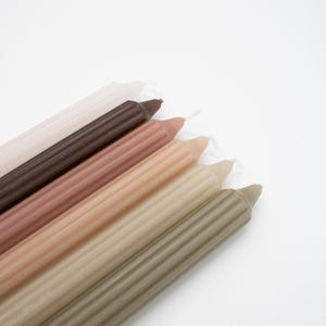 Könnte beinhalten: Ein Set mit sechs gerippten Kerzen in verschiedenen Brauntönen, Beige und Grün. Die Kerzen sind fächerförmig angeordnet, wobei die dunkelste Farbe oben und die hellste Farbe unten ist.