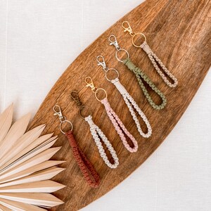 Könnte beinhalten: Sechs gehäkelte Schlüsselanhänger in verschiedenen Farben, darunter Braun, Rosa, Beige und Grün, mit goldenen und silbernen Schlüsselringen. Die Schlüsselanhänger werden auf einer Holzoberfläche präsentiert.