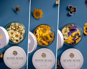 May include: Three glass jars filled with dried flowers. The jars are labelled "Rosie Rose" and have the text "Dekoration fur jeden Anlass". The flowers are white, yellow, and purple.