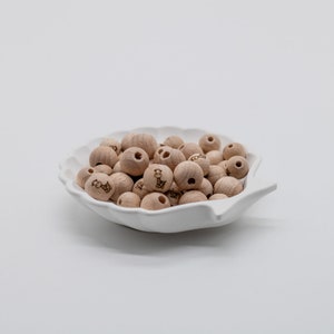 Puede incluir: Un cuenco de cerámica blanco lleno de cuentas de madera natural. Cada cuenta tiene un pequeño diseño de jirafa grabado.