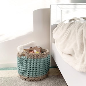 May include: A large, crocheted storage basket with a beige and teal color scheme. The basket is filled with folded blankets and books. The basket is sitting on a light gray rug in front of a white bed.